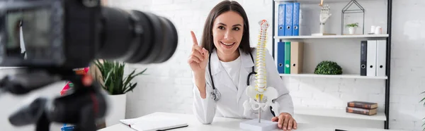 Médecin souriant gesticulant près du modèle de la colonne vertébrale et appareil photo numérique flou à la clinique, bannière — Photo de stock