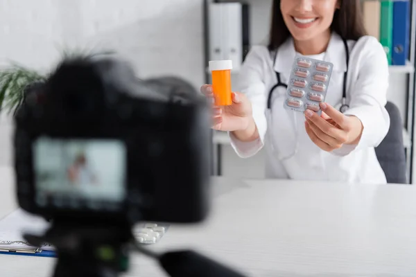 Vue recadrée d'un médecin souriant tenant des pilules près d'un appareil photo numérique flou dans une clinique — Photo de stock