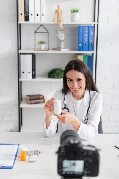 Médecin souriant tenant des pilules près du presse-papiers et de l'appareil photo numérique flou dans la clinique — Photo de stock