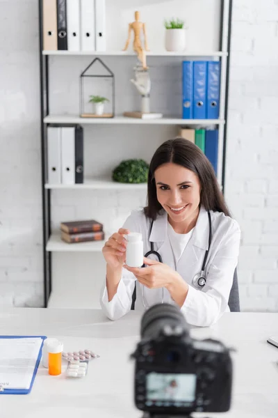 Médico sorrindo segurando pílulas perto da área de transferência e câmera digital borrada na clínica — Fotografia de Stock