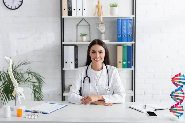 Lächelnder Arzt mit Blick auf Kamera in der Nähe von dna und Wirbelsäulenmodellen und Klemmbrett in Klinik — Stockfoto