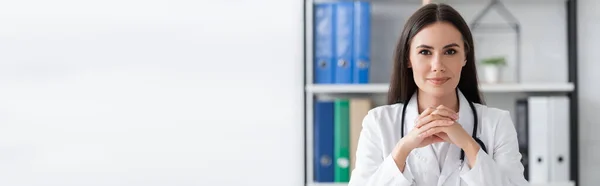Brunette médecin en manteau blanc regardant caméra à l'hôpital, bannière — Photo de stock