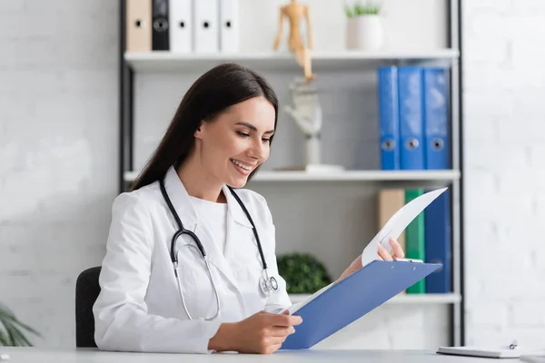 Lächelnder Arzt im weißen Kittel mit Klemmbrett in Klinik — Stockfoto