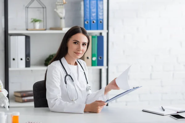 Doctor looking at camera while holding clipboard in clinic - foto de stock