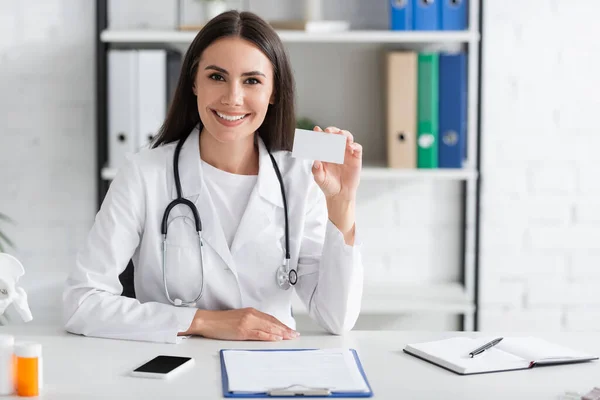 Médecin souriant tenant une carte de visite avec espace de copie près du smartphone et du presse-papiers dans la clinique — Photo de stock
