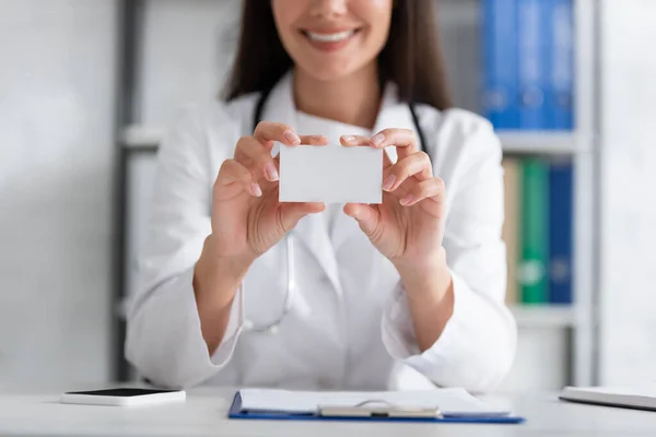 Cropped view of smiling blurred doctor holding empty business card near smartphone and clipboard in clinic — Stock Photo