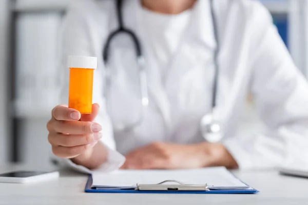 Vista recortada del médico borroso sosteniendo pastillas cerca del portapapeles y el teléfono inteligente en la clínica - foto de stock