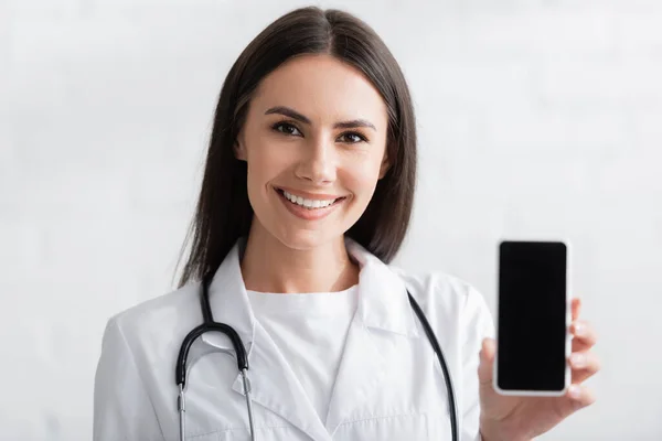 Happy brunette médecin tenant smartphone avec écran blanc — Photo de stock