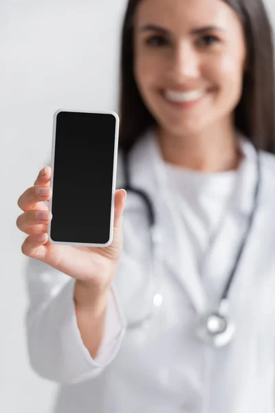 Médico borroso con bata blanca sosteniendo teléfono celular con pantalla en blanco en la clínica - foto de stock