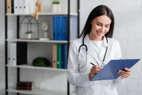 Fröhlicher Arzt schreibt im Krankenhaus auf Klemmbrett — Stockfoto