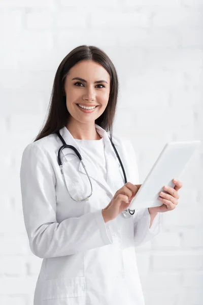 Brünette Ärztin lächelt in Kamera und benutzt digitales Tablet in Klinik — Stockfoto