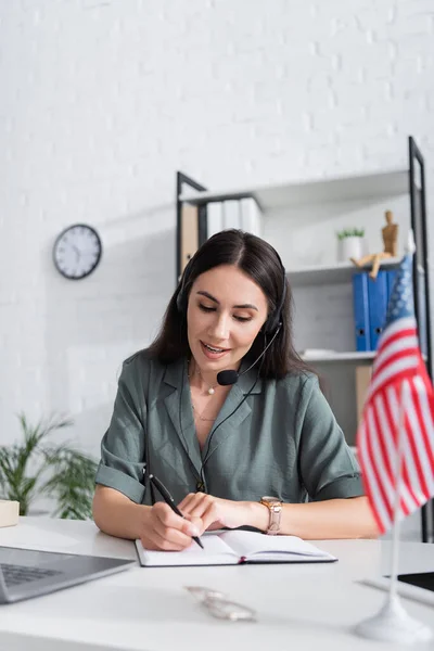 Smiling teacher in headset writing on notebook near american flag and laptop in school — Stock Photo