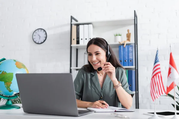 Positive teacher in headset having online lesson on laptop near globe and flags in school — Stock Photo