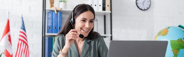 Smiling tutor in headset having video call on laptop near globe and flags in school, banner — Fotografia de Stock