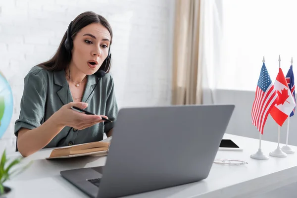 Teacher in headset talking near blurred laptop and flags on table in school — Stock Photo