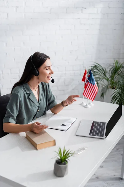Side view of smiling teacher in headset talking during video call on laptop near nook and flags in school — Stock Photo