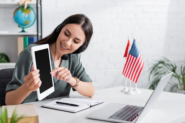 Smiling teacher in headset holding digital tablet during online lecture on laptop in school — Photo de stock