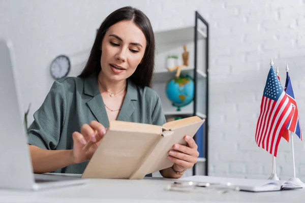 Teacher reading book during online lesson on laptop near flags in school — Stock Photo