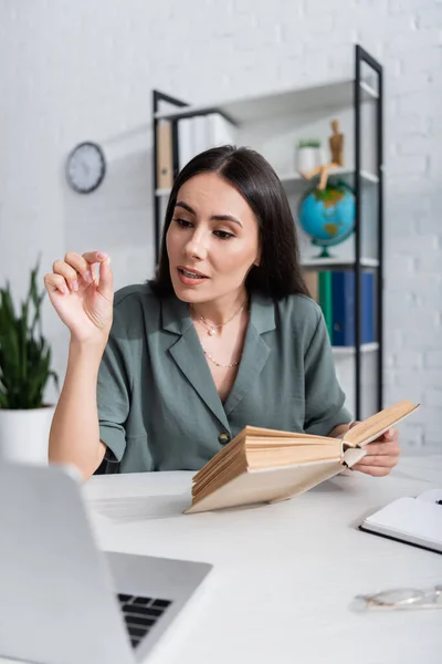 Lehrerin mit Buch spricht während Online-Unterricht am Laptop in der Schule — Stockfoto