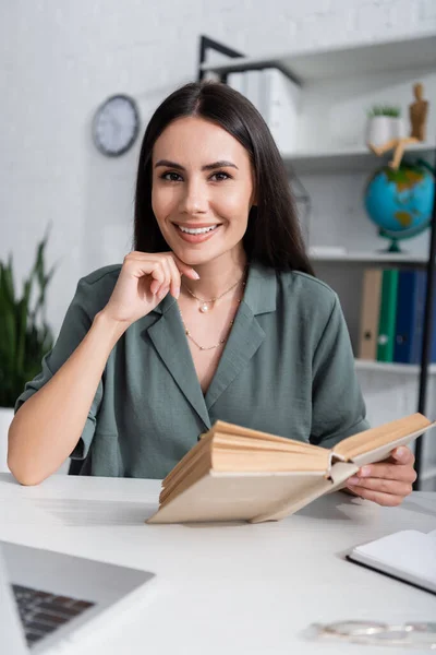 Lächelnder Lehrer hält Buch in der Hand und blickt in der Schule in die Kamera neben verschwommenem Laptop — Stockfoto