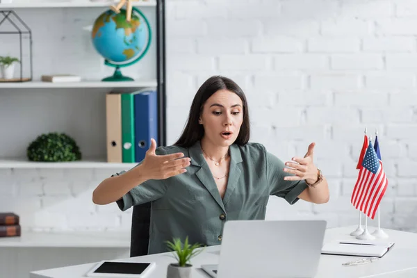 Teacher talking during video call on laptop near digital tablet and flags in school — Stockfoto