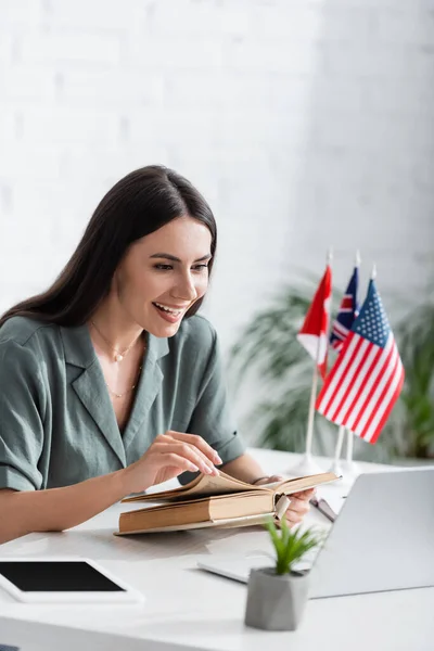 Cheerful teacher holding book and looking at laptop during online lecture in school — Foto stock