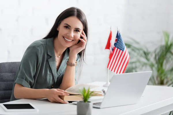 Positive teacher holding book near devices and flags in class — Stock Photo