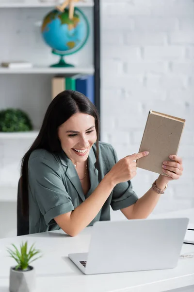 Positive teacher pointing at book during video call on laptop in school — Stockfoto