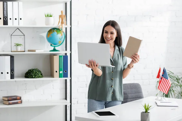 Insegnante positivo che tiene libro e laptop durante la lezione online a scuola — Foto stock