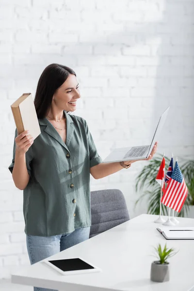 Fröhlicher Lehrer hält Buch und Laptop neben Fahnen und digitalem Tablet auf dem Tisch im Unterricht — Stockfoto