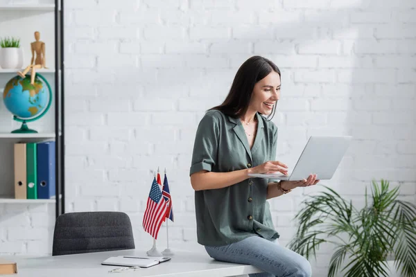 Cheerful teacher using laptop during online lecture near flags and notebook on table in school — Stock Photo