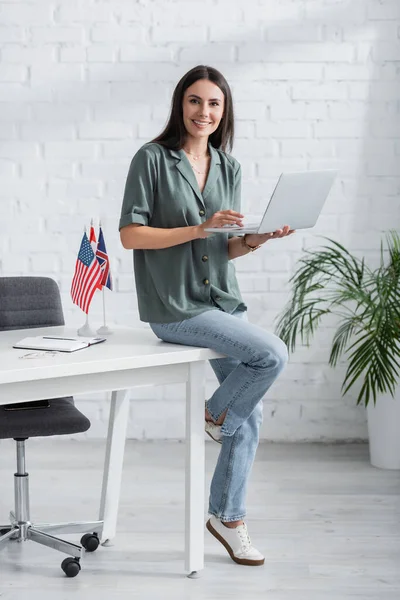 Smiling teacher holding laptop and looking at camera near flags on table in class — Stock Photo