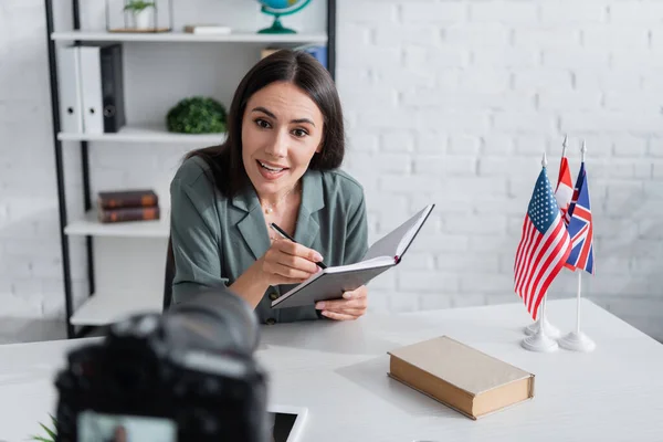 Smiling teacher holding notebook near book and blurred digital camera in class - foto de stock