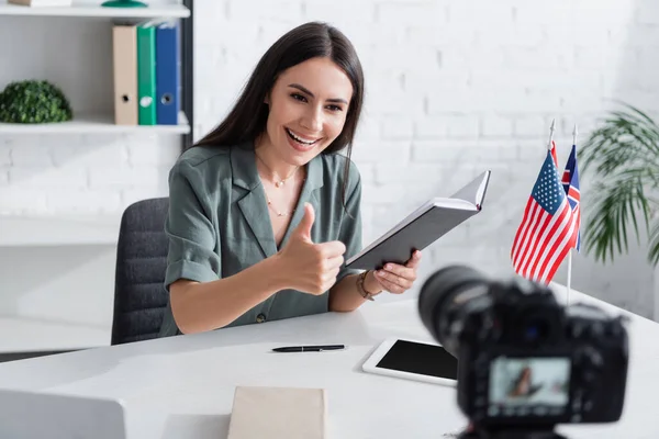 Lächelnder Lehrer zeigt Geste und hält Notizbuch in der Nähe von Digitalkamera im Unterricht — Stockfoto