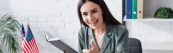 Positive teacher showing like and holding notebook near flags in school, banner — Stock Photo