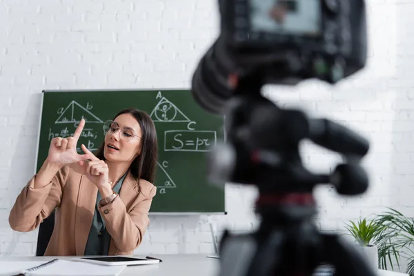 Profesor de gafas graduadas gesticulando cerca de pizarra con fórmulas matemáticas y cámara digital borrosa en clase - foto de stock