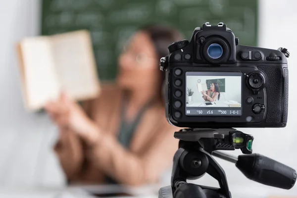 Profesor borroso sosteniendo libro cerca de la cámara digital en la escuela - foto de stock