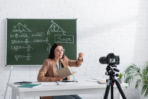 Tutor holding book near digital camera and chalkboard with mathematic formulas in class — Stock Photo
