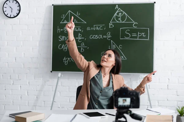 Professor sorridente apontando com os dedos perto de quadro-negro com fórmulas matemáticas e câmera digital na sala de aula — Fotografia de Stock