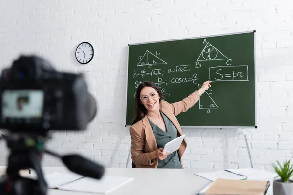 Glückliche Lehrerin hält digitales Tablet in der Hand und zeigt auf Mathe-Formeln auf der Tafel neben der Kamera in der Schule — Stockfoto
