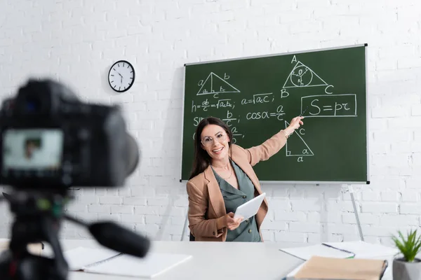 Professeur heureux tenant tablette numérique et pointant vers les formules mathématiques sur tableau près de la caméra à l'école — Photo de stock