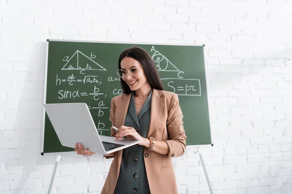 Enseignant souriant utilisant un ordinateur portable près des formules mathématiques sur tableau à l'école — Photo de stock