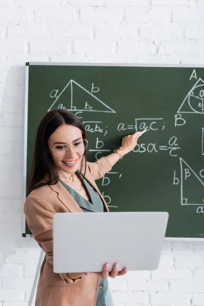 Profesor señalando fórmulas matemáticas en pizarra durante la lección en línea en clase - foto de stock