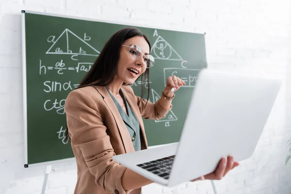 Вчитель розмовляє і тримає ноутбук під час онлайн-лекції біля дошки в класі — стокове фото