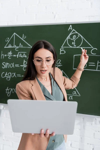 Profesor que tiene videollamada en el portátil cerca de pizarra con fórmulas matemáticas - foto de stock