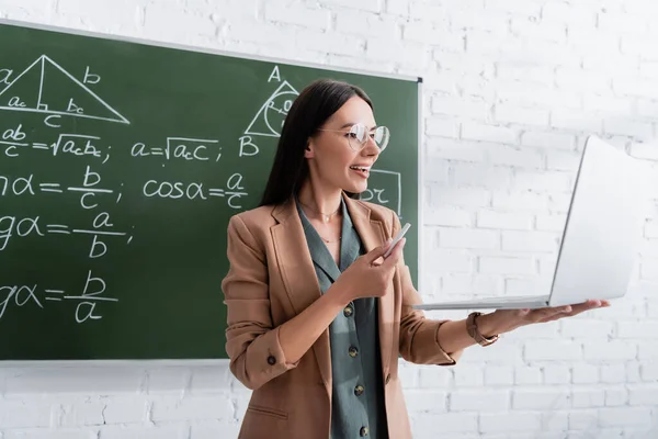Teacher having online lesson on laptop near math formulas on chalkboard — Stockfoto