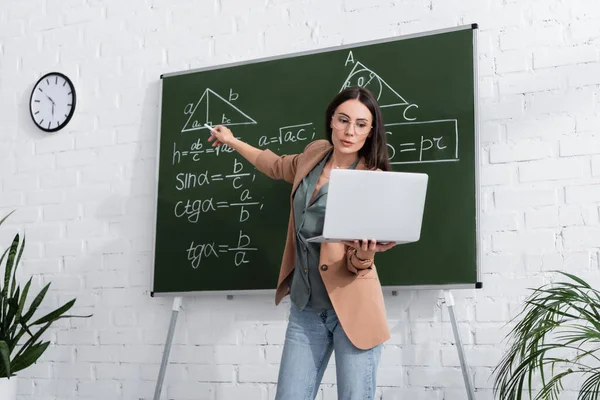 Profesora señalando fórmulas matemáticas en pizarra y sosteniendo la computadora portátil durante la lección en línea en el aula - foto de stock