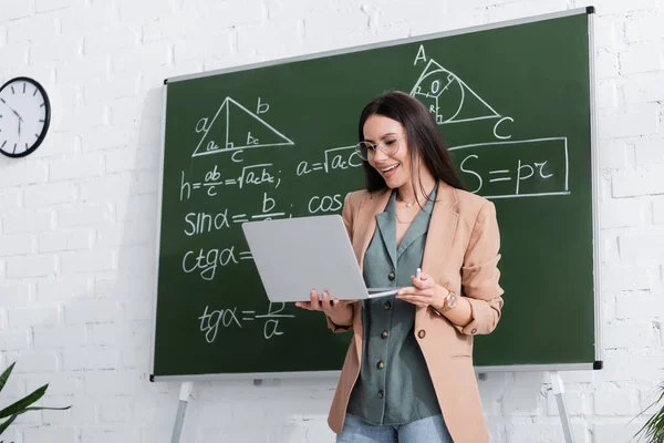 Positiver Lehrer hält Laptop während Online-Vorlesung neben Tafel mit Mathe-Formeln — Stockfoto