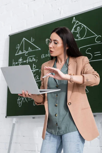 Teacher holding chalk and laptop during online lecture near chalkboard in classroom — Stock Photo