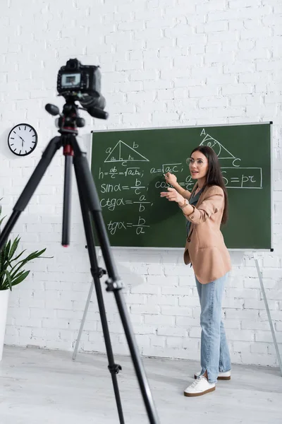 Teacher writing math formula on chalkboard near digital camera in class — Stock Photo